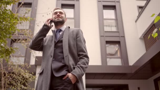Smiling handsome business man in coat talking by smartphone while standing outdoors in the city - Video