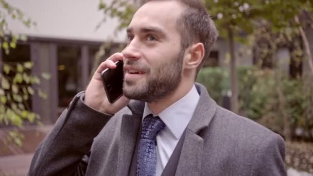 Happy handsome business man in coat talking by smartphone while standing outdoors in the city - Filmagem, Vídeo