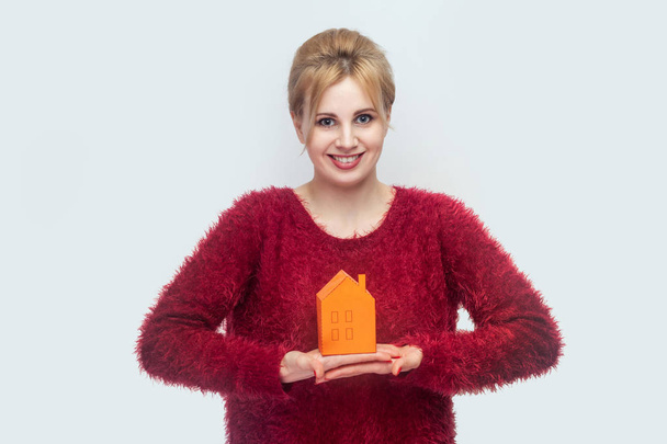 Cheerful positive optimistic young blonde woman in red sweater holding paper small house while planning new home to buy on light gray background - Fotografie, Obrázek
