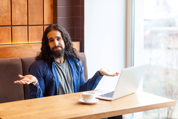 confused handsome young adult man freelancer in casual style sitting with raised hands in cafe near laptop and showing unknown gesture while looking at camera  - Foto, Imagen