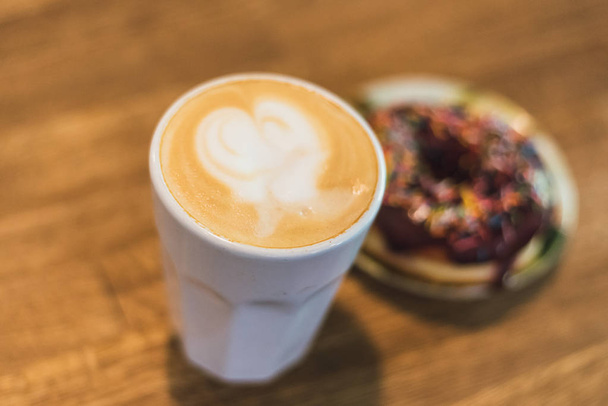 coffee with a heart made of milk and a donut on a wooden table. Coffee with a drawn heart and milk on a wooden table in a coffee shop. chocolate donut with scattering on the table next to the coffee - Photo, Image