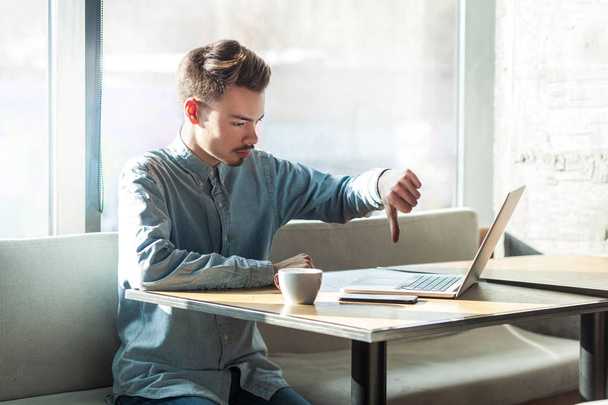 αρνητική κριτική μούσι νεαρό ελεύθερος σε μπλε πουκάμισο κάθεται στο καφέ και κάνοντας βιντεοκλήση στο laptop, ενώ δείχνοντας αντίχειρας προς τα κάτω στο σύντροφό του  - Φωτογραφία, εικόνα