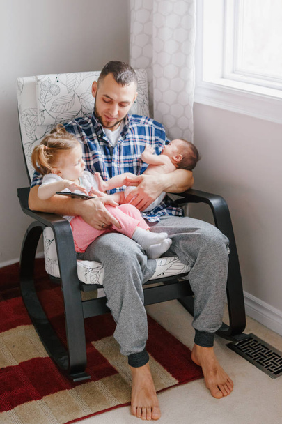 Kaukaski ojciec tata siedzi z córką malucha i noworodka syna oglądania bajki na smartphone. Szczery autentyczne życie rodzinne życia. Samotna opieka nad dziećmi w domu. - Zdjęcie, obraz