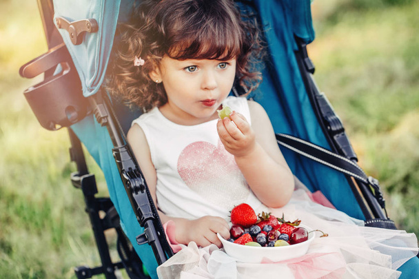 かわいいかわいい白人の幼児の赤ちゃんの女の子は外のベビーカーに座って果実を食べています。屋外で健康的なスナック食事をしている面白い子供。子供のための夏おいしいフィンガーフード. - 写真・画像