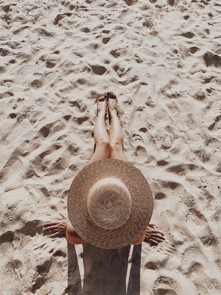 Summer travel fashion and vacation concept. Beautiful young, tanned woman with a straw hat is lying and relaxing on tropical beach with white sand showing legs. Top view, flat lay. - 写真・画像