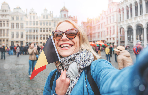 Женщина-туристка стоит с флагом Бельгии на фоне Гран-Пласа или Большой Рыночной площади в Брюсселе
 - Фото, изображение