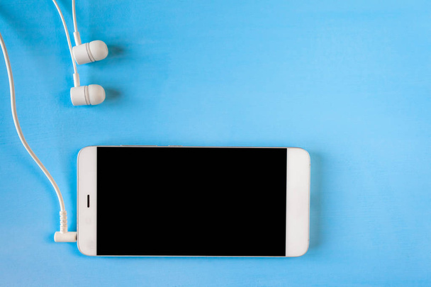 white smartphone with connected white headphones on blue background, copyspace - Photo, Image