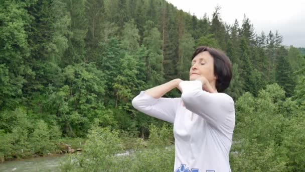 Starsza kobieta z krótkimi ciemnymi włosami robi ćwiczenia rozciągające dla relaksu rano poza krajobrazem lasu i gór - Materiał filmowy, wideo