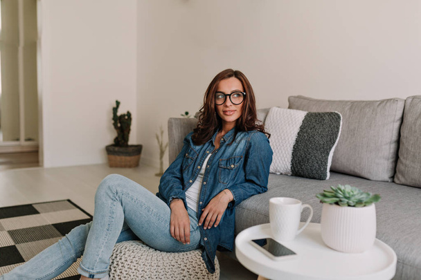 Foto mulher relaxante em casa e olhando para o lado com grande sorriso vestindo camisa jeans e óculos
.  - Foto, Imagem