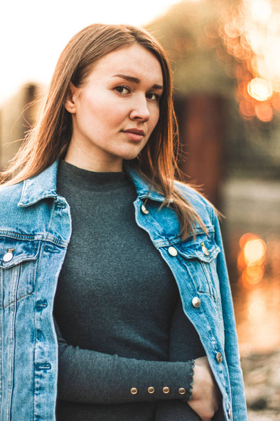 Portrait of the attractive, slender, beautiful young Caucasian blonde girl in a jeans jacket. Smiling girl enjoys fine warm sunner weather against the forest - Photo, Image