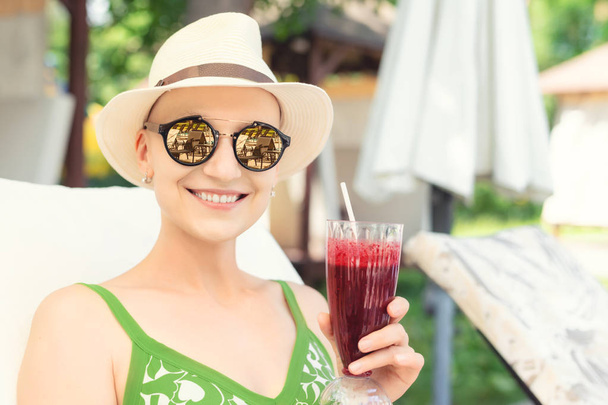 Young happy woman holding fresh berry smoothie cocktail enjoying vacation at resort on hot sunny day. young caucasian bald woman in hat and swimsuite enjoy life after surviving breast cancer - Photo, Image