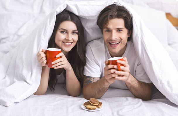 Χαρούμενο ζευγάρι που κρύβεται κάτω από πάπλωμα με φλιτζάνια και μπισκότα - Φωτογραφία, εικόνα