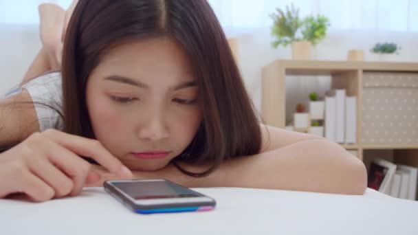 Mladá asijská žena, která používá smartphone a kontroluje společenské sdělovací prostředky, se šťastně usmívá, když leží na posteli, když se ráno probudila, krásná atraktivní dívka usmívající se relaxace v ložnici doma. - Záběry, video