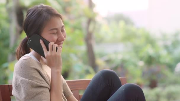 Mujer asiática usando teléfono móvil hablando con amigos, mujer relajarse sintiéndose feliz sentado en la mesa en el jardín por la mañana. Estilo de vida de las mujeres relajarse en el hogar concepto
. - Imágenes, Vídeo