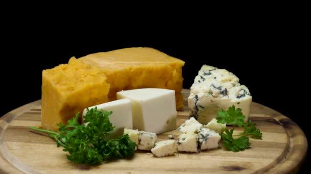 Close up for french delicious aged cheeses choped and served on wooden board isolated on black background. Frame. Cheddar, parmesan, and blue pieces of cheese with parsley. - Footage, Video