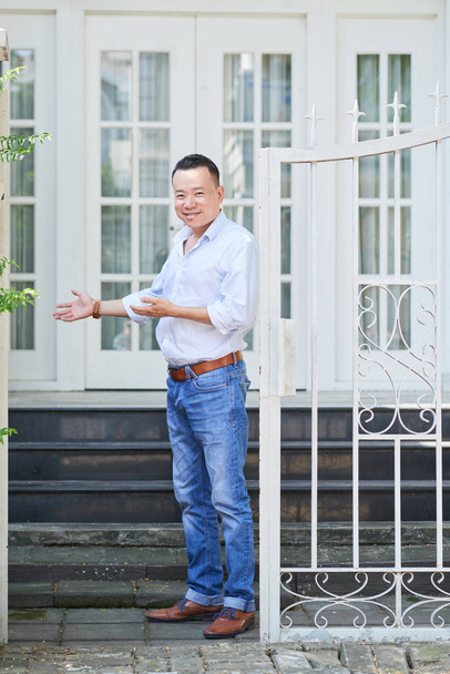 カジュアルな服を着たアジア人男性の肖像画が門の近くに立ち、あなたを彼の家に招待する  - 写真・画像