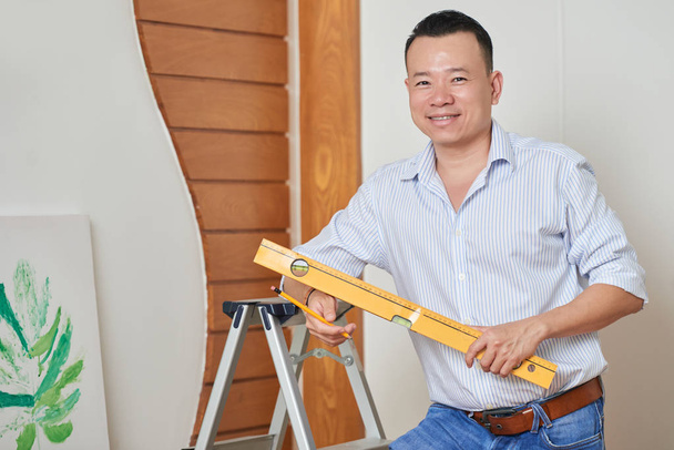 Portrait of young smiling man holding level and pencil and standing near the wall, he is doing repairment himself - Photo, Image