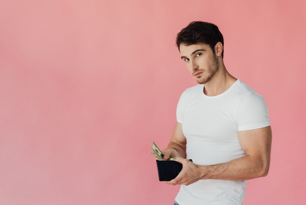 muscular man in white t-shirt holding wallet with dollar banknotes and looking at camera isolated on pink - Fotografie, Obrázek