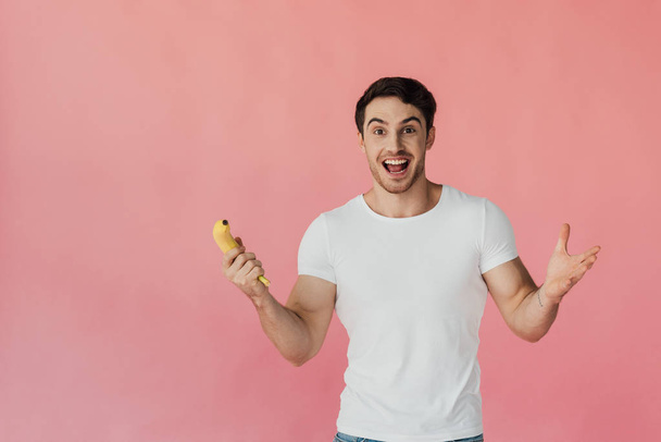 front view of excited muscular man in white t-shirt holding banana isolated on pink - Фото, зображення
