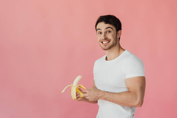 smiling muscular man in white t-shirt holding banana and looking at camera isolated on pink - Foto, Bild