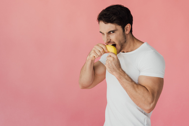 hungry muscular man in white t-shirt eating banana isolated on pink - Foto, Imagem