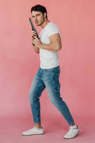 повний вигляд м'язистого чоловіка в білій футболці тримає пістолет ізольовано на рожевому
 - Фото, зображення