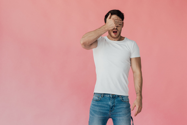 vista frontal del hombre musculoso conmocionado en camiseta blanca que cubre los ojos con la mano aislada en rosa
 - Foto, imagen