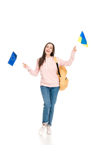 full length view of smiling student with backpack holding Ukrainian and European flags isolated on white - 写真・画像
