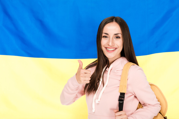 sorridente bruna giovane donna con zaino mostrando pollice su sfondo bandiera ucraina
 - Foto, immagini