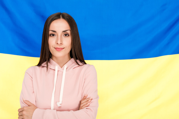 glimlachend brunette jonge vrouw met gekruiste armen staande op Oekraïense vlag achtergrond - Foto, afbeelding