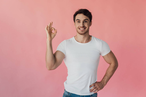 front view of smiling muscular man in white t-shirt showing okay sign isolated on pink - Valokuva, kuva