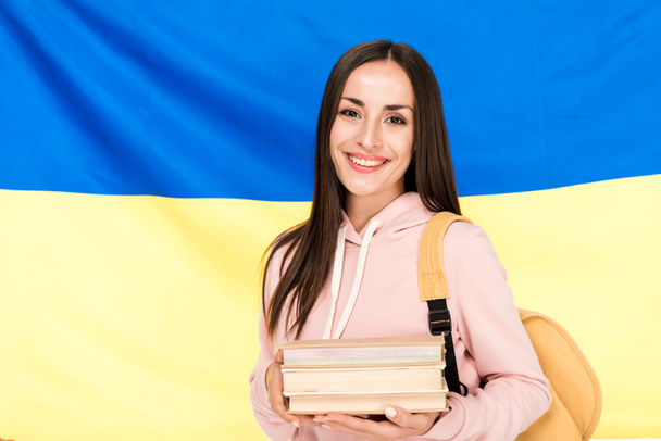 glimlachend brunette jonge vrouw met rugzak en boeken over Oekraïense vlag achtergrond - Foto, afbeelding