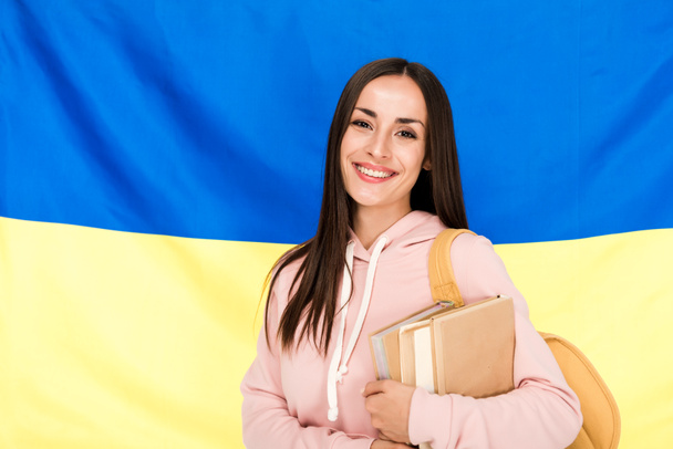 Gelukkig brunette jonge vrouw met rugzak en boeken over Oekraïense vlag achtergrond - Foto, afbeelding