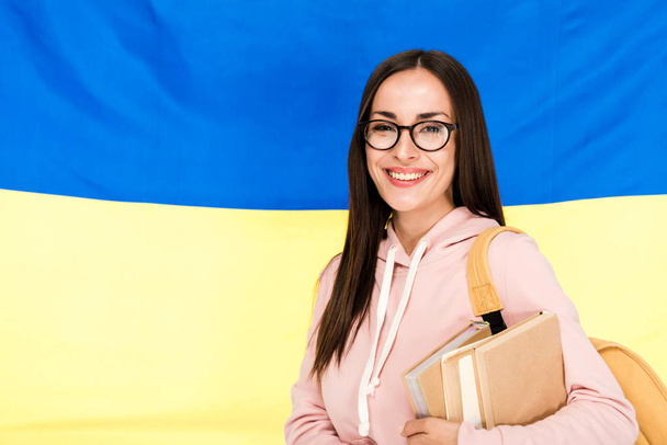 Lächelnde brünette junge Frau in Brille mit Rucksack und Büchern auf ukrainischem Fahnenhintergrund - Foto, Bild