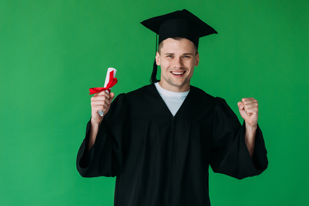smiling student in academic cap holding diploma with red ribbon and showing yes gesture isolated on green - Фото, изображение