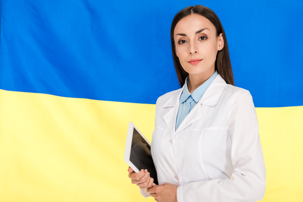 врач в белом халате держит планшет на фоне украинского флага
 - Фото, изображение