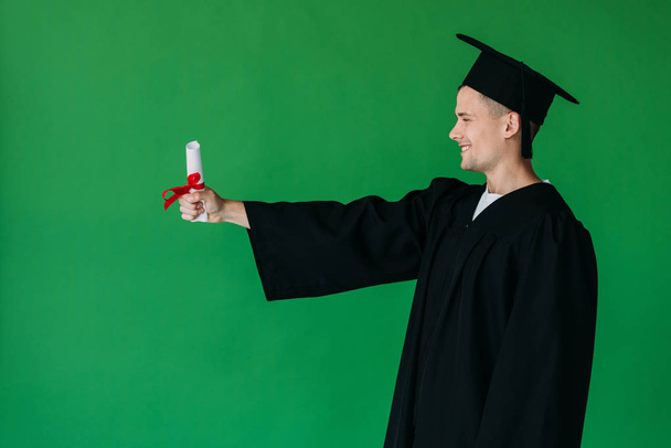 vista lateral del estudiante sonriente en la gorra académica sosteniendo diploma con cinta roja aislada en verde
 - Foto, imagen