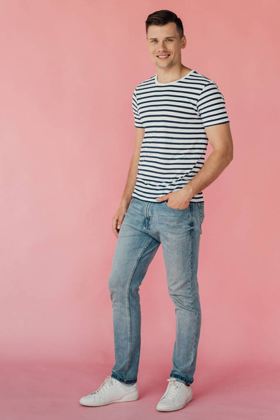 full length view of smiling man in jeans and striped t-shirt with hand in pocket looking at camera on pink - Fotó, kép