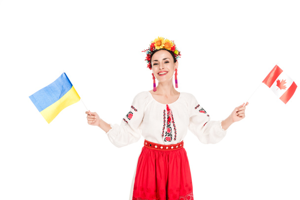 sonriente morena joven en traje nacional ucraniano sosteniendo banderas de Ucrania y Canadá aislado en blanco
 - Foto, Imagen