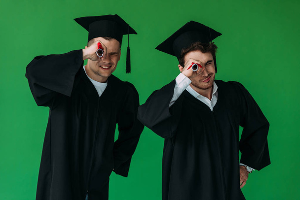 dois estudantes felizes em gorros acadêmicos que possuem diplomas isolados em verde
 - Foto, Imagem