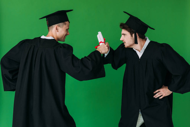 two students in academic caps holding diplomas and looking at each other isolated on green - Photo, Image