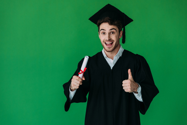 excited student in academic cap holding diploma with red ribbon and showing thumb up isolated on green - 写真・画像