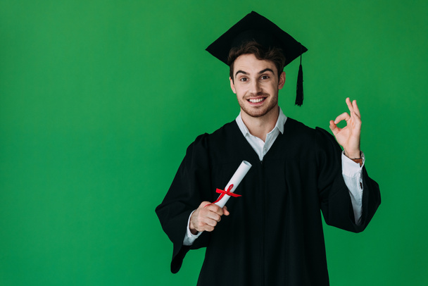vista frontal del estudiante en el diploma de la celebración de la gorra académica con cinta roja y mostrando signo bien aislado en verde
 - Foto, Imagen