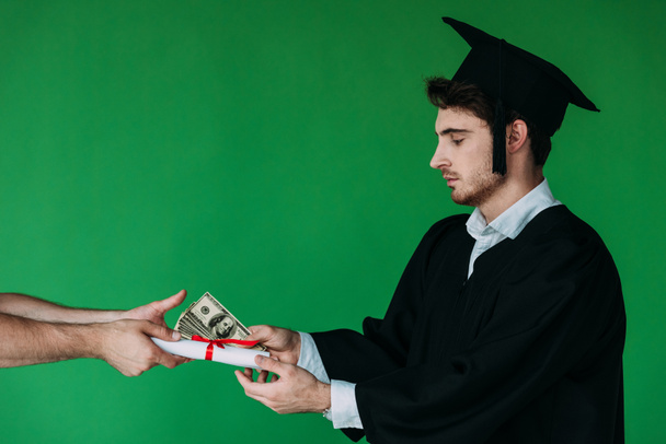 student in academic cap holding diploma with red ribbon and offering bribe isolated on green - Photo, Image