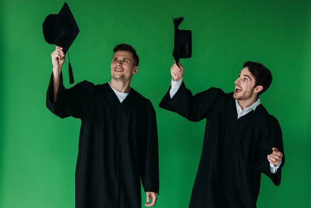 two excited students holding academic caps isolated on green - Foto, afbeelding