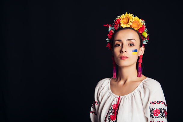 brunette jonge vrouw in nationale Oekraïense kostuum en bloemen krans kijken naar camera geïsoleerd op zwart - Foto, afbeelding