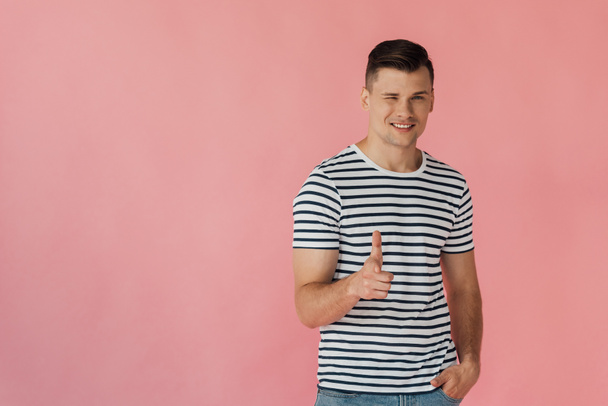 smiling man in striped t-shirt standing with hand in pocket, blinking and showing thumb up isolated on pink - Фото, зображення