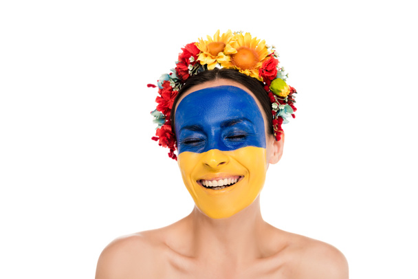 nahá mladá žena v květinové věnce s namalovanou ukrajinskou vlajkou na kůži usmívající se s zavřenýma očima izolovanými na bílém - Fotografie, Obrázek