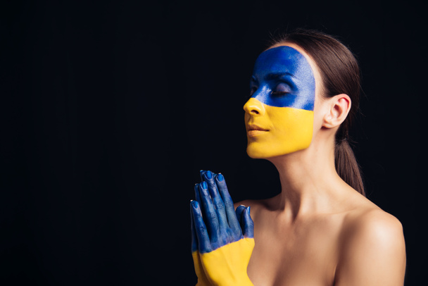 naked young woman with painted Ukrainian flag on skin praying with closed eyes isolated on black - Фото, зображення