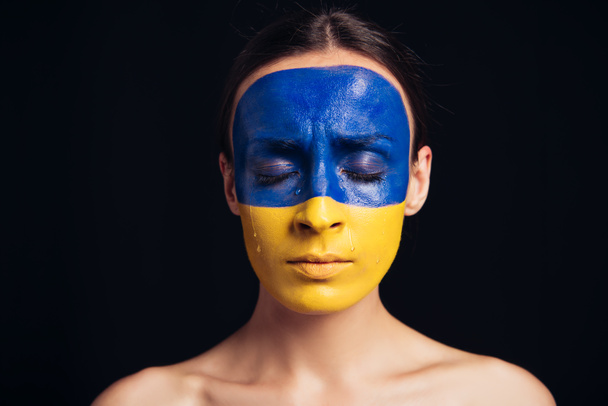 mujer joven desnuda con bandera ucraniana pintada en la piel y los ojos cerrados aislados en negro
 - Foto, Imagen